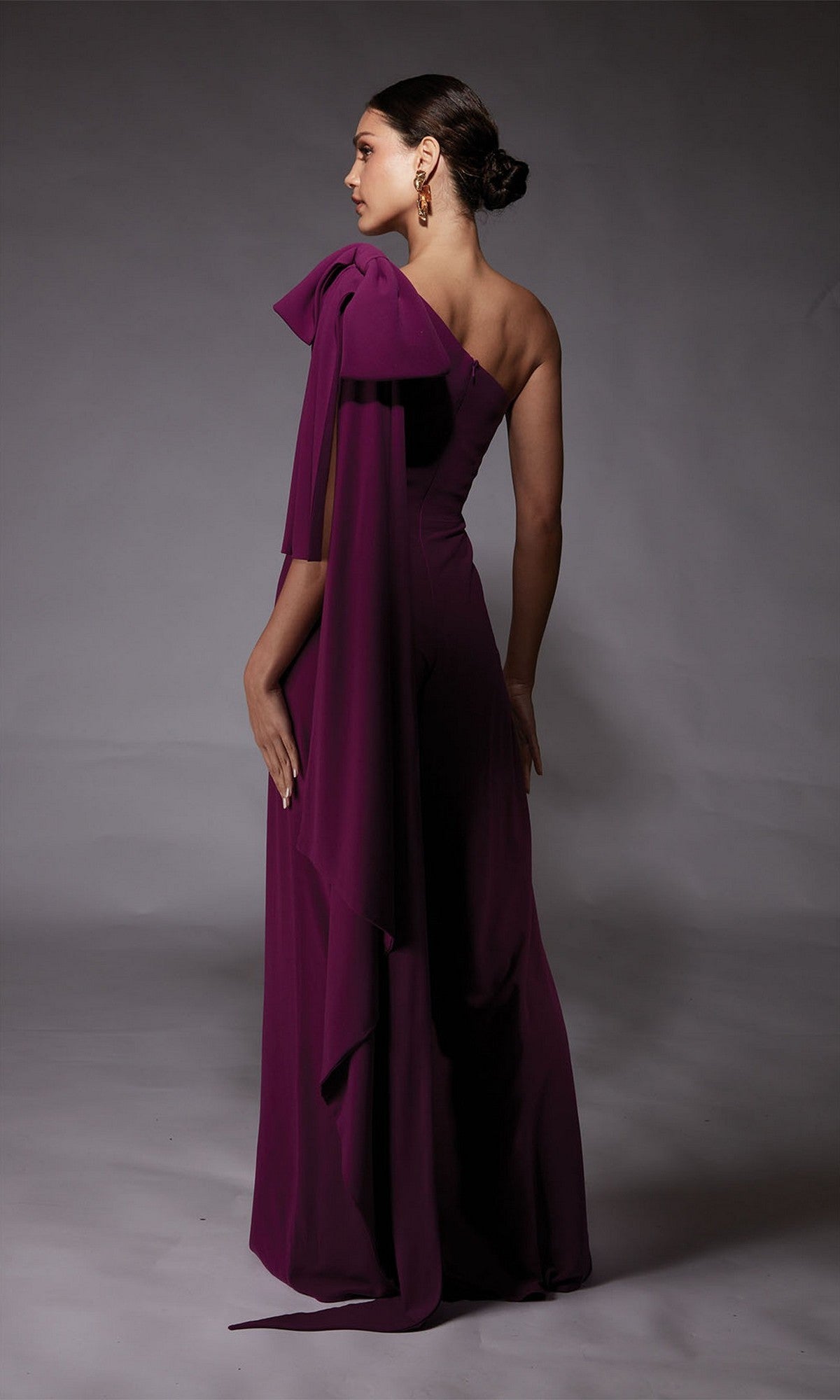 A woman stands in a studio facing away, wearing the Alyce 70065 long formal one-shoulder jumpsuit with draped sleeve detail—a striking alternative to a cocktail dress.